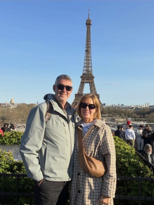 Two people wearing sunglasses pose for a photograph in front of the Eiffel Tower in Paris. The image is taken from a popular viewing area, with hedges, visitors and the Paris skyline visible in the background beneath a bright blue sky.