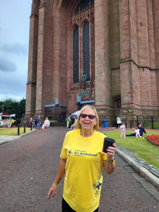 A North West Air Ambulance Charity supporter wearing a yellow charity T-shirt stands outside Liverpool Cathedral holding a takeaway coffee cup. The cathedral's large stone entrance towers in the background, with visitors visible around the grounds.