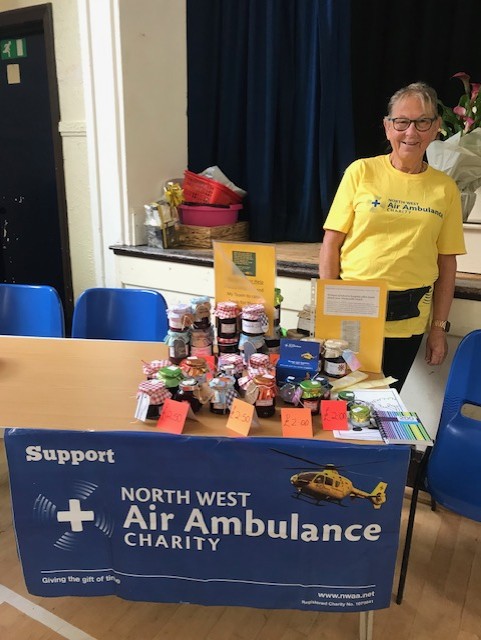 A North West Air Ambulance Charity supporter stands beside a fundraising stall indoors. The table is covered with a charity banner and displays jars of preserves, small gifts and fundraising materials, with price labels visible on several items.